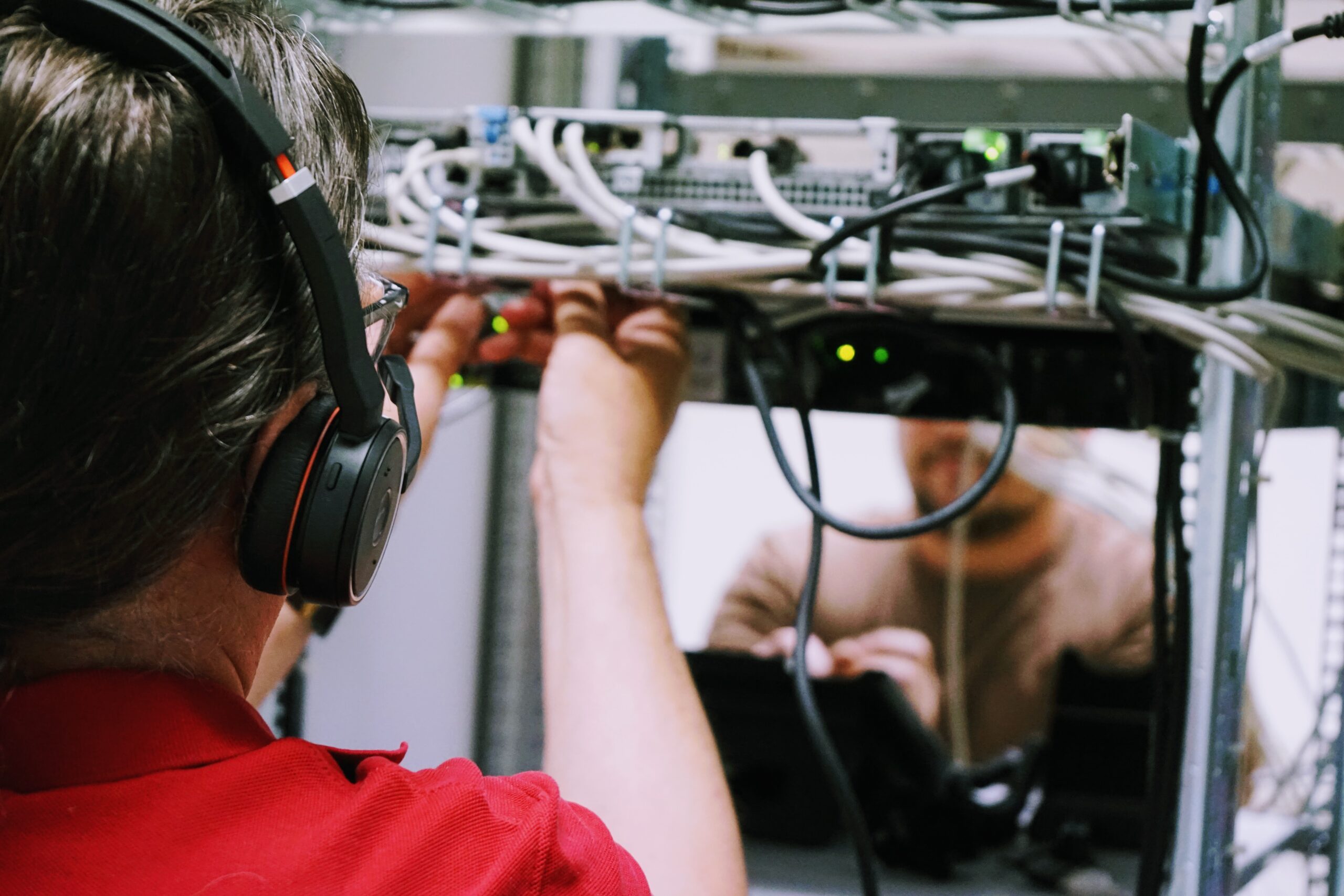 Zu sehen sind zwei Techniker an den Technikschränken eines Kernnetzstandortes: Links im Bild die Person ist von hinten zu sehen, sie trägt Kopfhörer und hat die Hände an Kabeln. Rechts im Bild die Person gegenüber ist von vorne zusehen, aber nur der untere Teil des Gesichts (mit Bart).