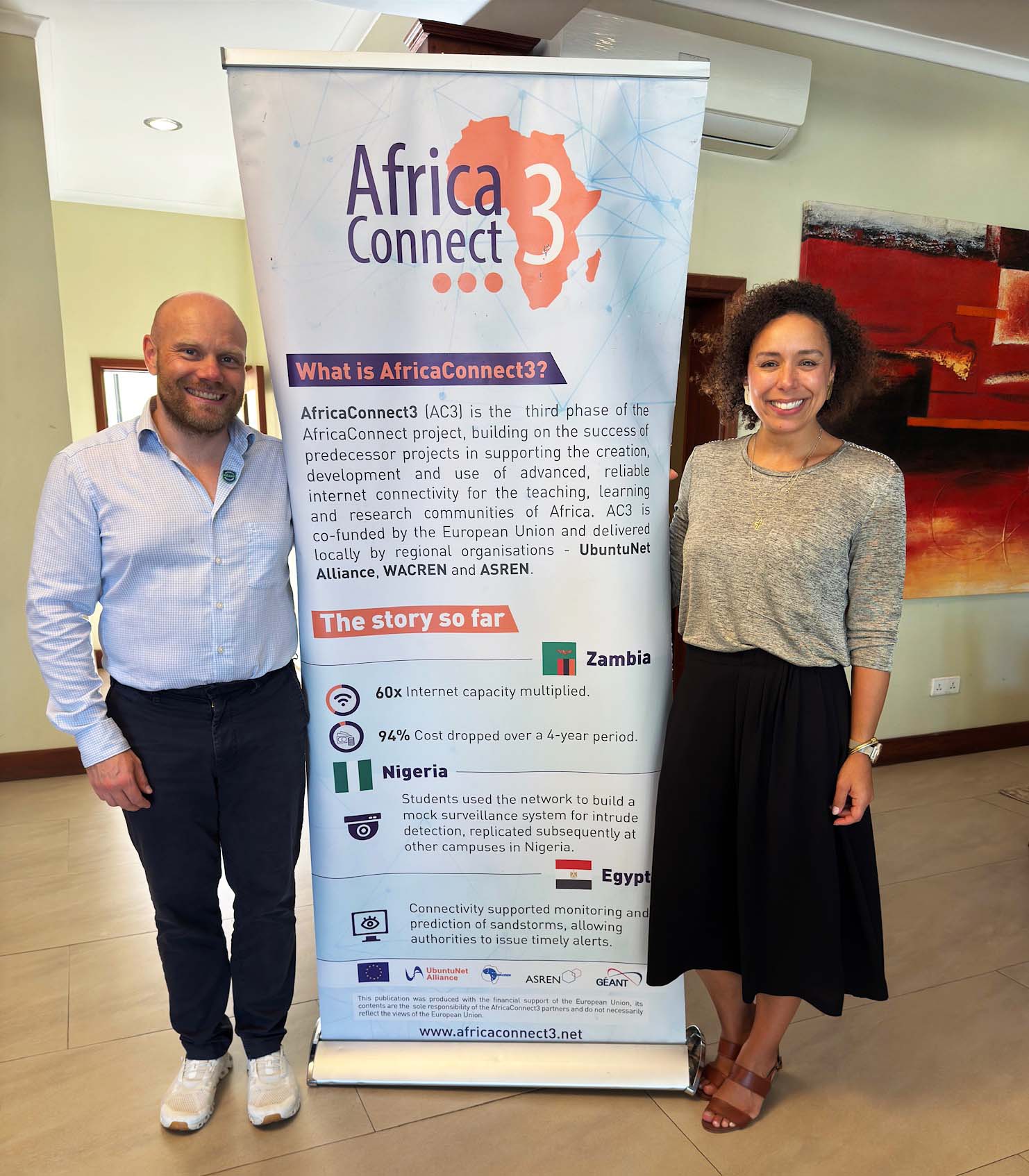 Im Bild zu sehen sind Trainerin Ana Alves (rechts) und Trainer Michel Gerdes (links) vom GÉANT-Security-Bootcamp bei ihrem Aufenthalt in Tansania. Zwischen ihnen ist ein Aufsteller mit Informationen zum EU-Projekt AfricaConnect3. Foto: GÉANT
