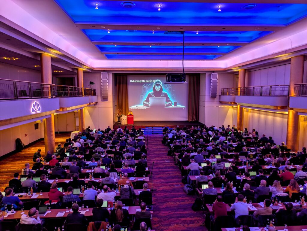 Weitwinkelaufnahme eines großen Konferenzsaals mit vielen Teilnehmenden, die an langen Tischen sitzen und einem Vortrag folgen. Die Beleuchtung im Raum ist überwiegend blau-violett. Auf der Leinwand ist eine Präsentation mit dem Titel „Cyberangriffe durch KI“ zu sehen, darunter ein Bild eines anonymen, maskierten Hackers mit Laptop. Eine Person steht vorne am Rednerpult und hält den Vortrag.