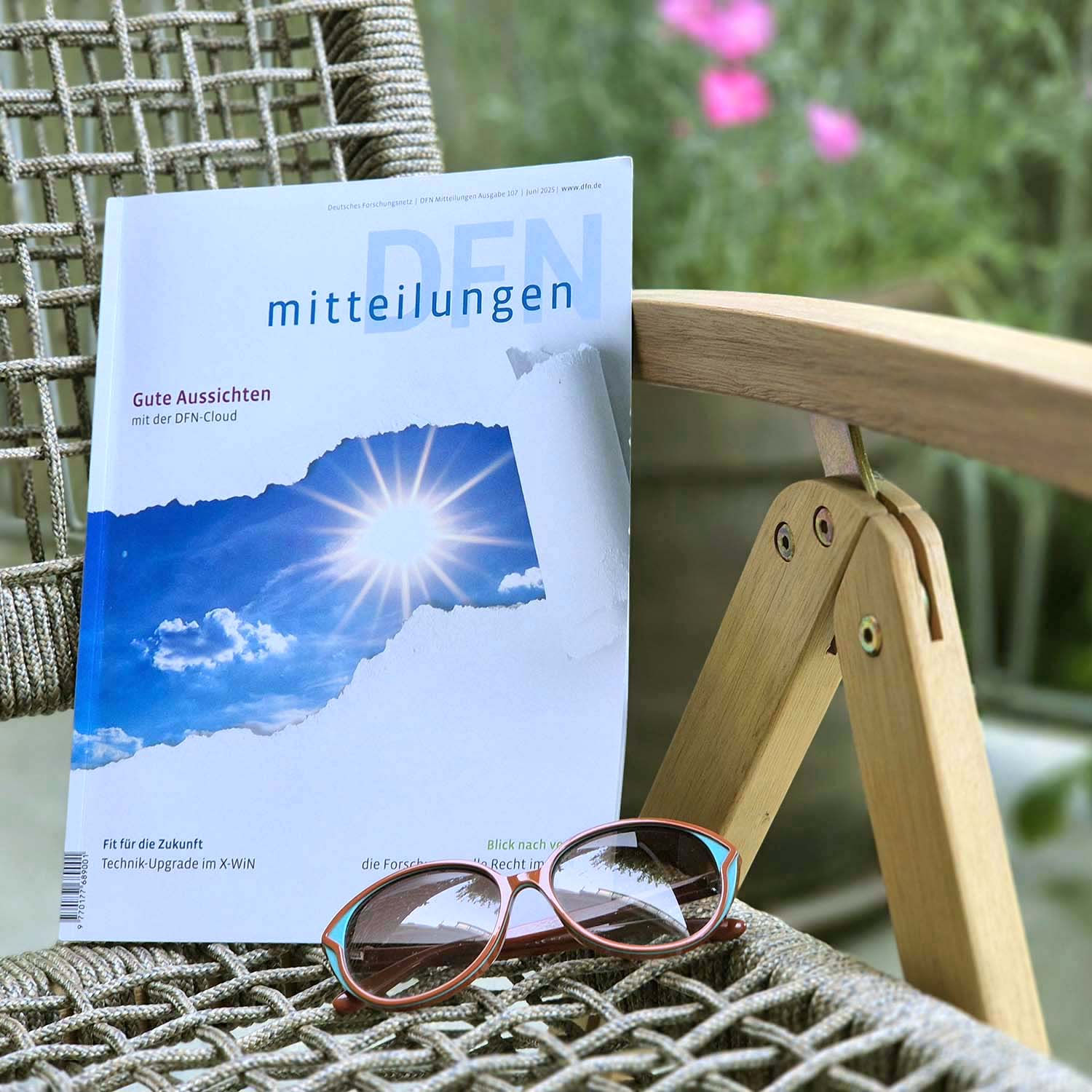 Ein Exemplar der Zeitschrift „DFN-Mitteilungen“ liegt auf einem Gartenstuhl mit Holz- und Seilbespannung. Der Titel der Ausgabe lautet: „Gute Aussichten mit der DFN-Cloud“. Auf dem Cover ist ein Sonnenbild zu sehen, das wie durch aufgerissenes Papier sichtbar wird. Auf dem Stuhl liegt auch eine rot-türkise Sonnenbrille. Im Hintergrund unscharf: grüne Pflanzen und pinke Blüten – sommerliche Atmosphäre. Foto: DFN-Verein