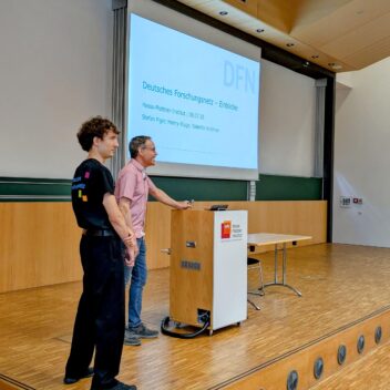 Zwei Männer stehen in einem modernen Hörsaal des Hasso-Plattner-Instituts auf einer kleinen Bühne neben einem Rednerpult. Der Jüngere trägt ein schwarzes T-Shirt mit bunter Aufschrift auf dem Rücken, der ältere ein rosa Hemd. Im Hintergrund ist eine große Präsentationsfolie zu sehen mit dem Titel „Deutsches Forschungsnetz – Einblicke“ und den Namen der Vortragenden: Stefan Piger, Henry Kluge, Valentin Kirchner. Die Präsentation fand am 08.07.2025 statt. Der Raum ist mit Holz verkleidet, mit grüner Tafelwand und großen Lautsprechern an der Decke.