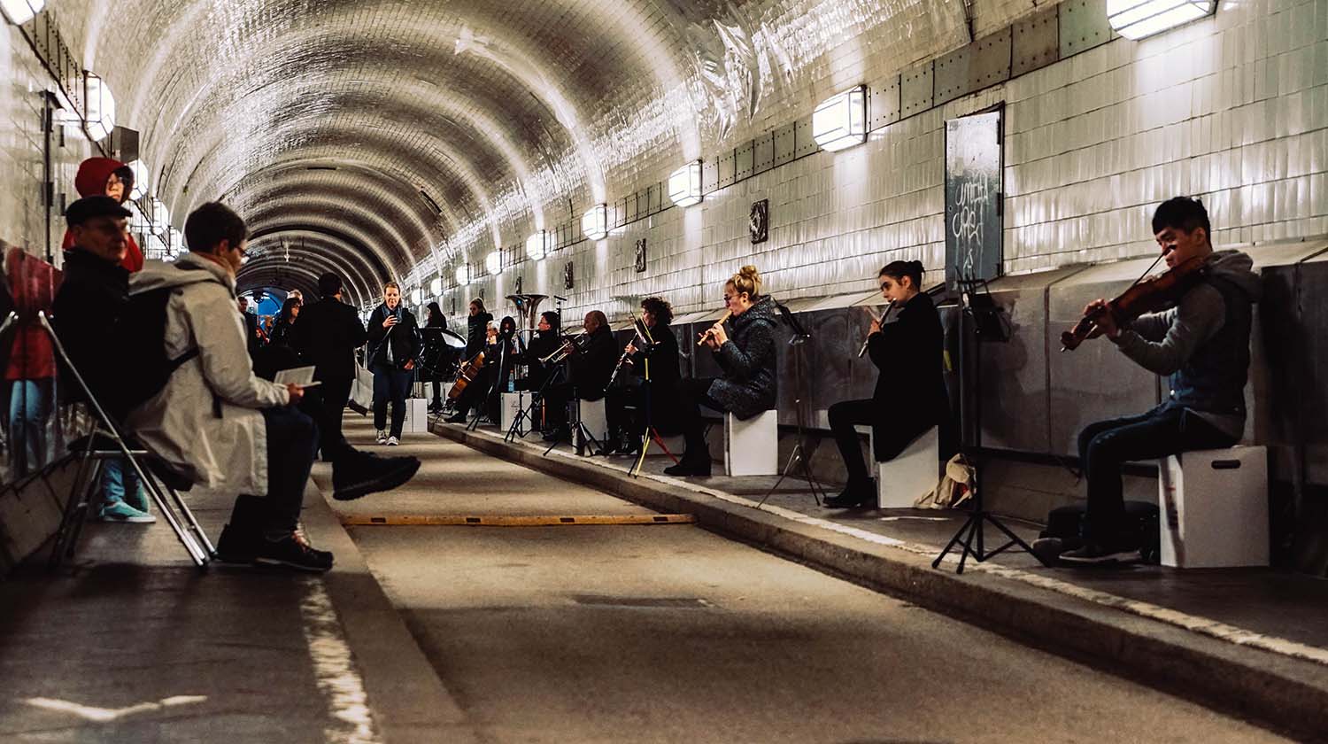 Musicians perform a classical concert inside a tunnel with white tiled walls and arched ceiling. The ensemble, including flutists, violinists, and cellists, is seated along the tunnel wall while a small audience listens from the opposite side. The unique underground setting creates an intimate and atmospheric performance space.