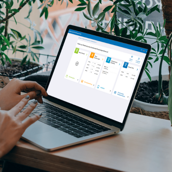 Person arbeitet an einem Laptop, auf dem ein Dashboard mit mehreren farbigen Karten und Informationen zu verschiedenen Diensten angezeigt wird. Der Laptop steht auf einem Holztisch neben mehreren Zimmerpflanzen