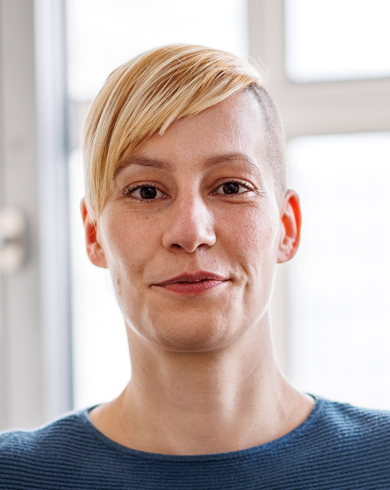 Porträt einer Frau mit kurz rasierten Haaren auf einer Seite und blondem, seitlich fallendem Pony. Sie trägt ein blaues Oberteil und blickt mit neutralem Gesichtsausdruck direkt in die Kamera. Der Hintergrund ist unscharf und zeigt ein Fenster mit Tageslicht.