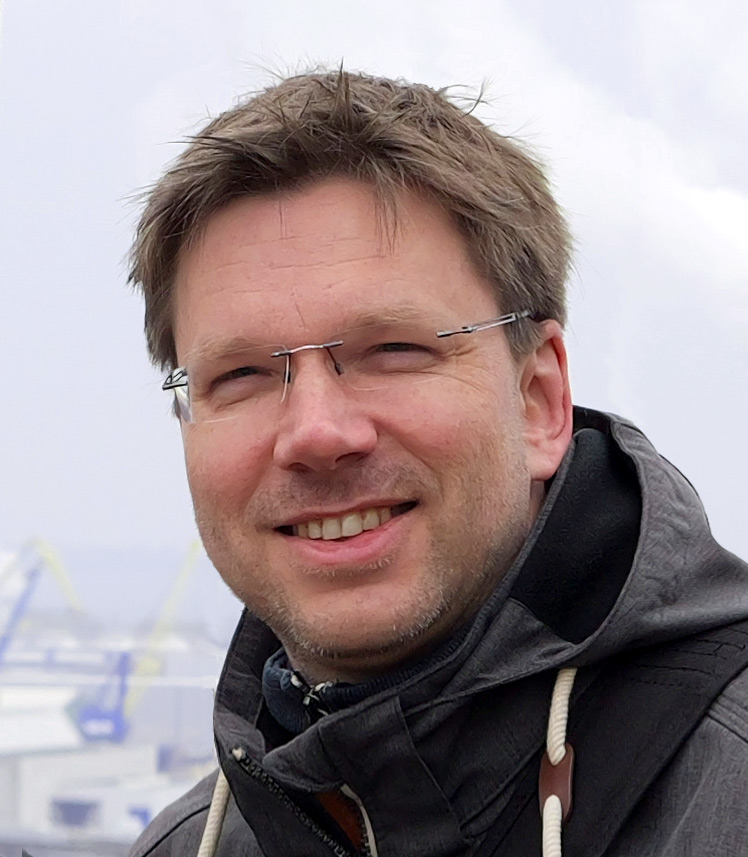 Porträt eines Mannes mit Brille und kurzem, braunem Haar. Er trägt eine dunkelgraue Outdoorjacke mit weißen Kordeln und lächelt in die Kamera. Im Hintergrund sind unscharf Hafenkräne und Schiffe zu erkennen, was auf eine maritime Umgebung hinweist.