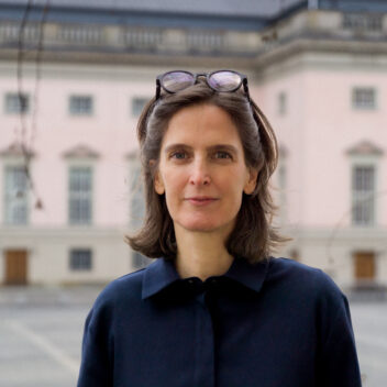 Eine Frau mittleren Alters mit schulterlangem braunem Haar steht im Freien vor einem historischen Gebäude mit rosa Fassade und hohen Fenstern. Sie trägt eine dunkelblaue Bluse und hat eine Brille auf dem Kopf. Die Szene wirkt ruhig und der Hintergrund ist leicht unscharf, sodass der Fokus auf der Frau liegt.