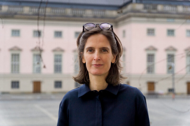 Eine Frau mittleren Alters mit schulterlangem braunem Haar steht im Freien vor einem historischen Gebäude mit rosa Fassade und hohen Fenstern. Sie trägt eine dunkelblaue Bluse und hat eine Brille auf dem Kopf. Die Szene wirkt ruhig und der Hintergrund ist leicht unscharf, sodass der Fokus auf der Frau liegt.