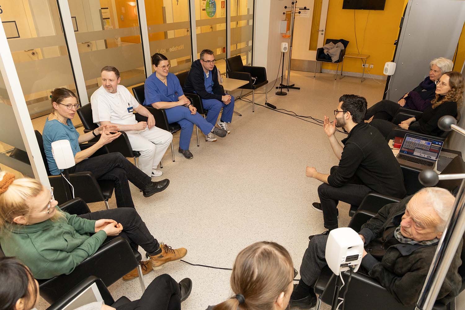 A group of healthcare professionals, researchers, and artists sit in a circle in a hospital hallway, engaged in a discussion. Some wear medical uniforms, while others have laptops or sound equipment, indicating an interdisciplinary meeting, likely related to a health and music project.