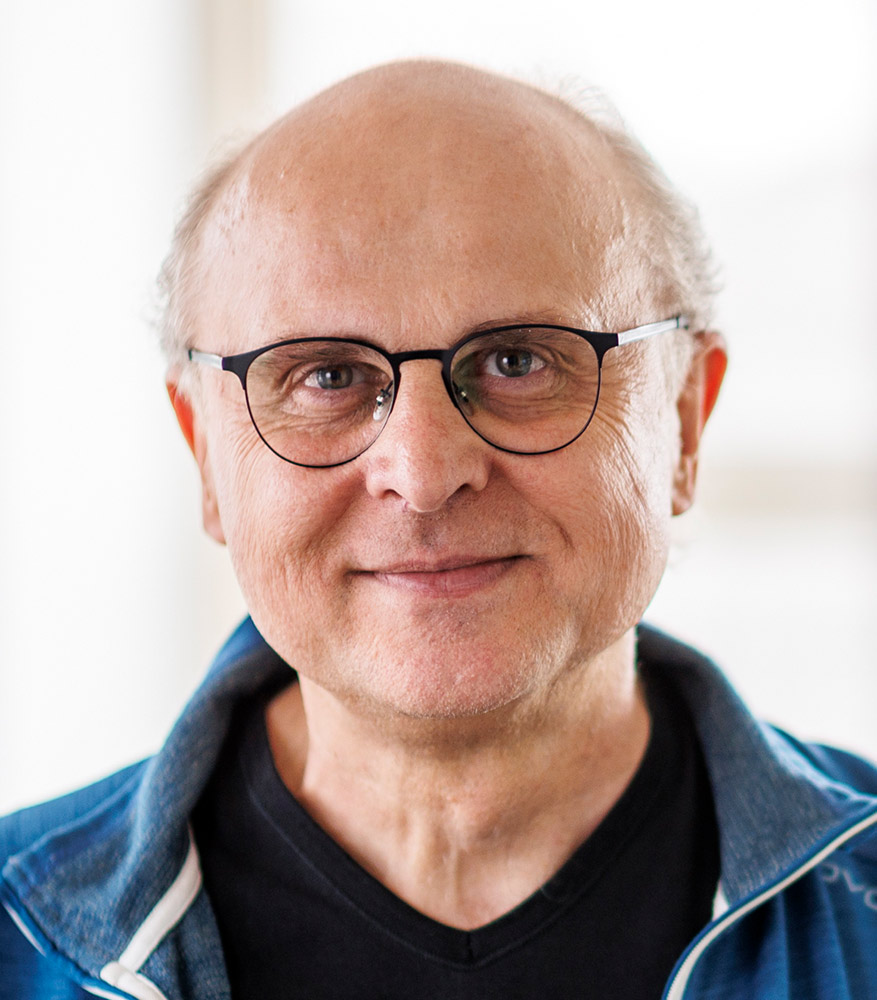 Ein älterer Mann mit Brille und kurzem, lichtem Haar lächelt freundlich in die Kamera. Er trägt eine schwarze T-Shirt und eine blau-graue Jacke mit Reißverschluss. Der Hintergrund ist weich und hell, was den Fokus auf sein Gesicht legt.