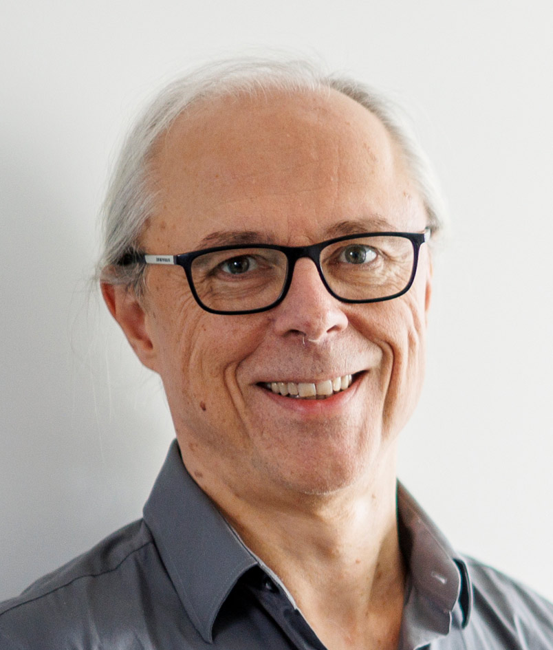 Porträt eines älteren Mannes mit langen, grauen Haaren, die nach hinten gebunden sind. Er trägt eine dunkle Brille und ein graues Hemd. Der Mann lächelt freundlich, der Hintergrund ist neutral weiß.