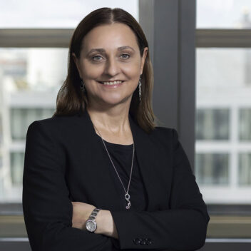 Eine Frau mittleren Alters mit braunen, schulterlangen Haaren steht mit verschränkten Armen vor einem Fenster mit Blick auf moderne Gebäude. Sie trägt ein schwarzes Outfit, eine silberne Uhr, eine lange Halskette mit Anhänger und passende Ohrringe. Sie lächelt selbstbewusst in die Kamera.