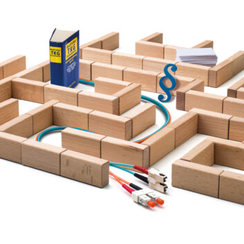 Ein Labyrinth aus Holzklötzen symbolisiert ein komplexes System. In dem Labyrinth verlaufen bunte Netzwerkkabel, ein Gesetzbuch mit der Aufschrift ‚TKG‘ (Telekommunikationsgesetz), ein blauer Paragrafenzeichen und ein Stapel Papier. Das Bild steht sinnbildlich für die regulatorischen und technischen Hürden in der Telekommunikation.
