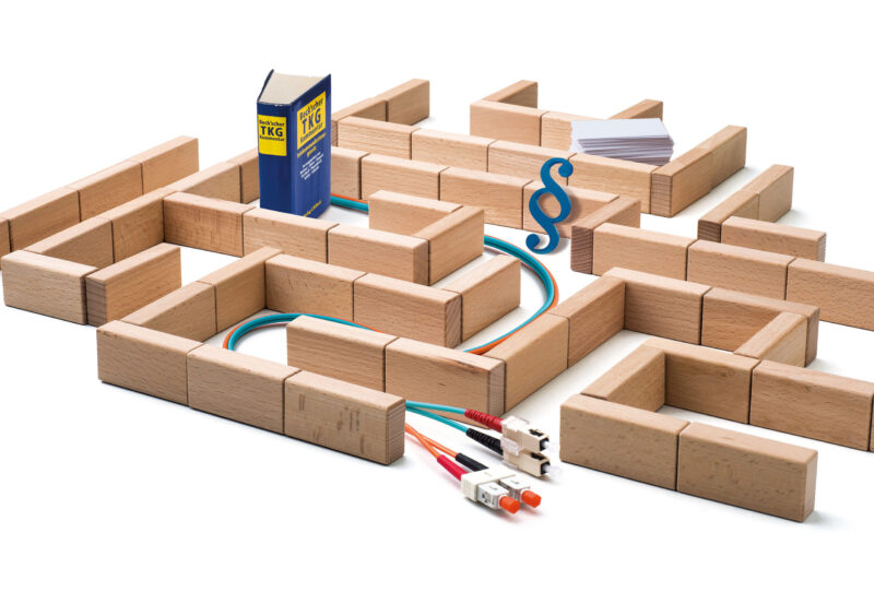 Ein Labyrinth aus Holzklötzen symbolisiert ein komplexes System. In dem Labyrinth verlaufen bunte Netzwerkkabel, ein Gesetzbuch mit der Aufschrift ‚TKG‘ (Telekommunikationsgesetz), ein blauer Paragrafenzeichen und ein Stapel Papier. Das Bild steht sinnbildlich für die regulatorischen und technischen Hürden in der Telekommunikation.