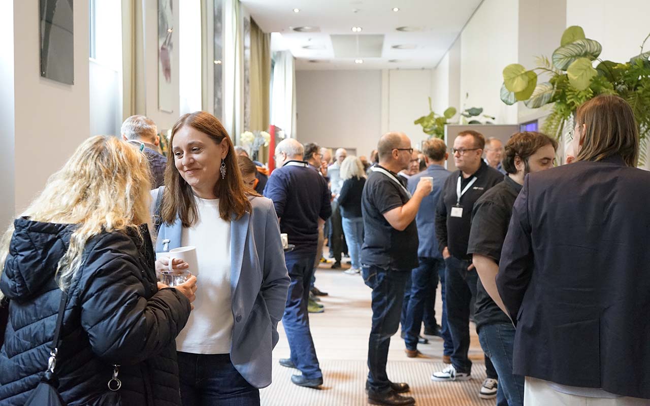 Menschen unterhalten sich in einem hellen Konferenzflur während einer Veranstaltung oder Kaffeepause. Zwei Frauen im Vordergrund führen ein Gespräch, während im Hintergrund mehrere Gruppen von Personen stehen und sich ebenfalls austauschen. Viele Teilnehmer tragen Namensschilder. Die Atmosphäre wirkt freundlich und professionell.