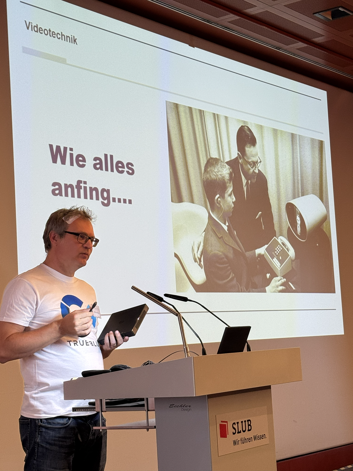 Ein Vortragender steht an einem Rednerpult mit der Aufschrift „SLUB – Wir führen Wissen.“ und hält ein kleines Gerät in der Hand. Im Hintergrund wird eine Präsentationsfolie mit dem Titel „Videotechnik“ gezeigt. Auf der Folie steht „Wie alles anfing…“ und es ist ein historisches Schwarz-Weiß-Foto von zwei Männern zu sehen, die ein frühes Videokommunikationssystem benutzen.