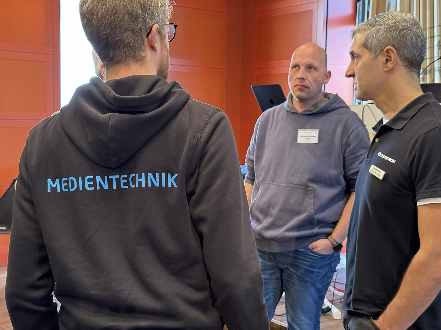 Drei Männer stehen in einem Raum mit orangefarbenen Wänden und unterhalten sich. Einer von ihnen trägt eine schwarze Jacke mit der blauen Aufschrift „MEDIENTECHNIK“ auf dem Rücken. Die beiden anderen tragen Namensschilder und dunkle Oberteile. Im Hintergrund sind technische Geräte und ein Laptop zu sehen.