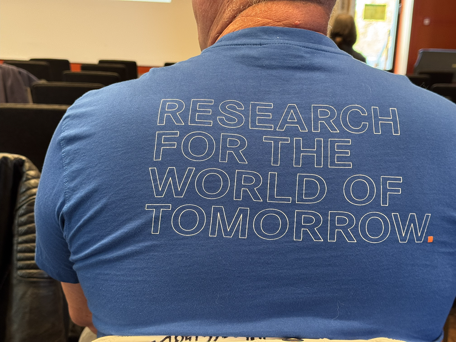 Ein Mann sitzt in einem Vortragssaal und trägt ein blaues T-Shirt mit der weißen Aufschrift „RESEARCH FOR THE WORLD OF TOMORROW“. Im Hintergrund sind einige weitere Personen und Stuhlreihen zu sehen.