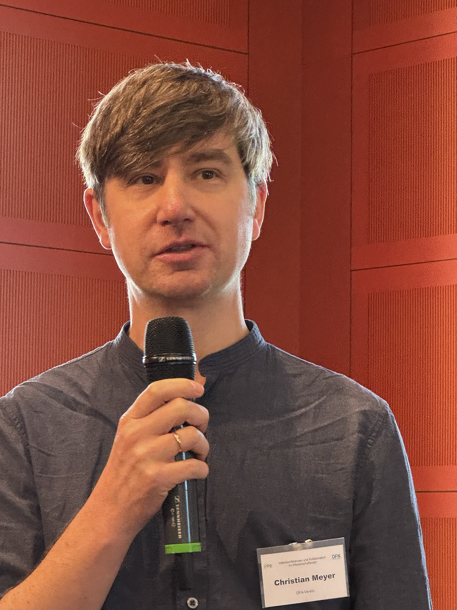 ChatGPT:Eine Person steht vor einer roten Wand und spricht in ein Mikrofon. Sie trägt ein dunkelblaues Hemd und ein Namensschild mit der Aufschrift „Christian Meyer“.