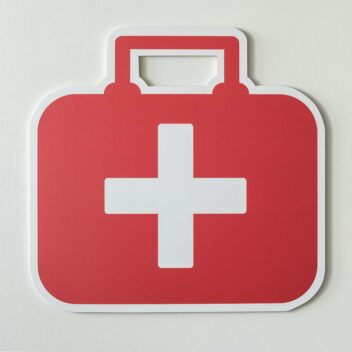 Rotes Erste-Hilfe-Symbol in Form eines roten Koffers mit weißem Kreuz auf der Vorderseite aus Pappe, vor einem hellen Hintergrund.