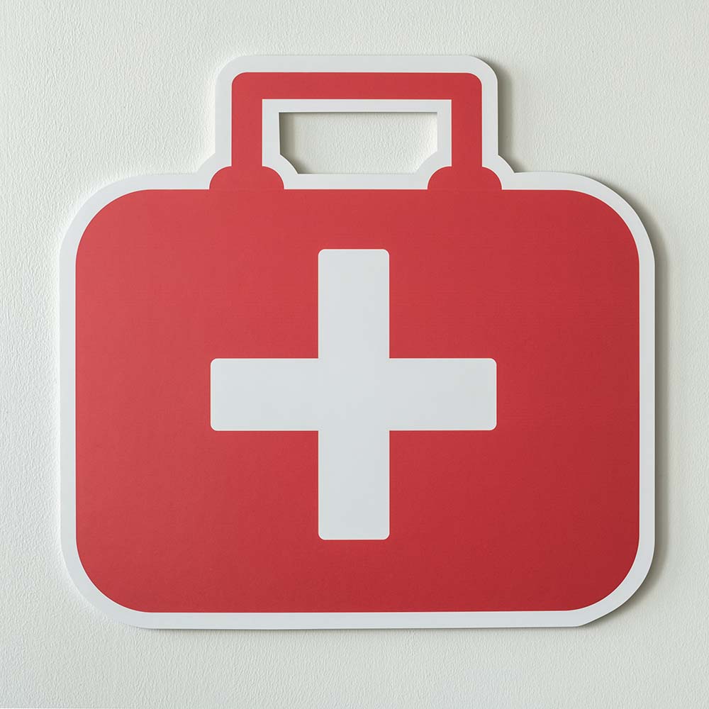 Rotes Erste-Hilfe-Symbol in Form eines roten Koffers mit weißem Kreuz auf der Vorderseite aus Pappe, vor einem hellen Hintergrund.