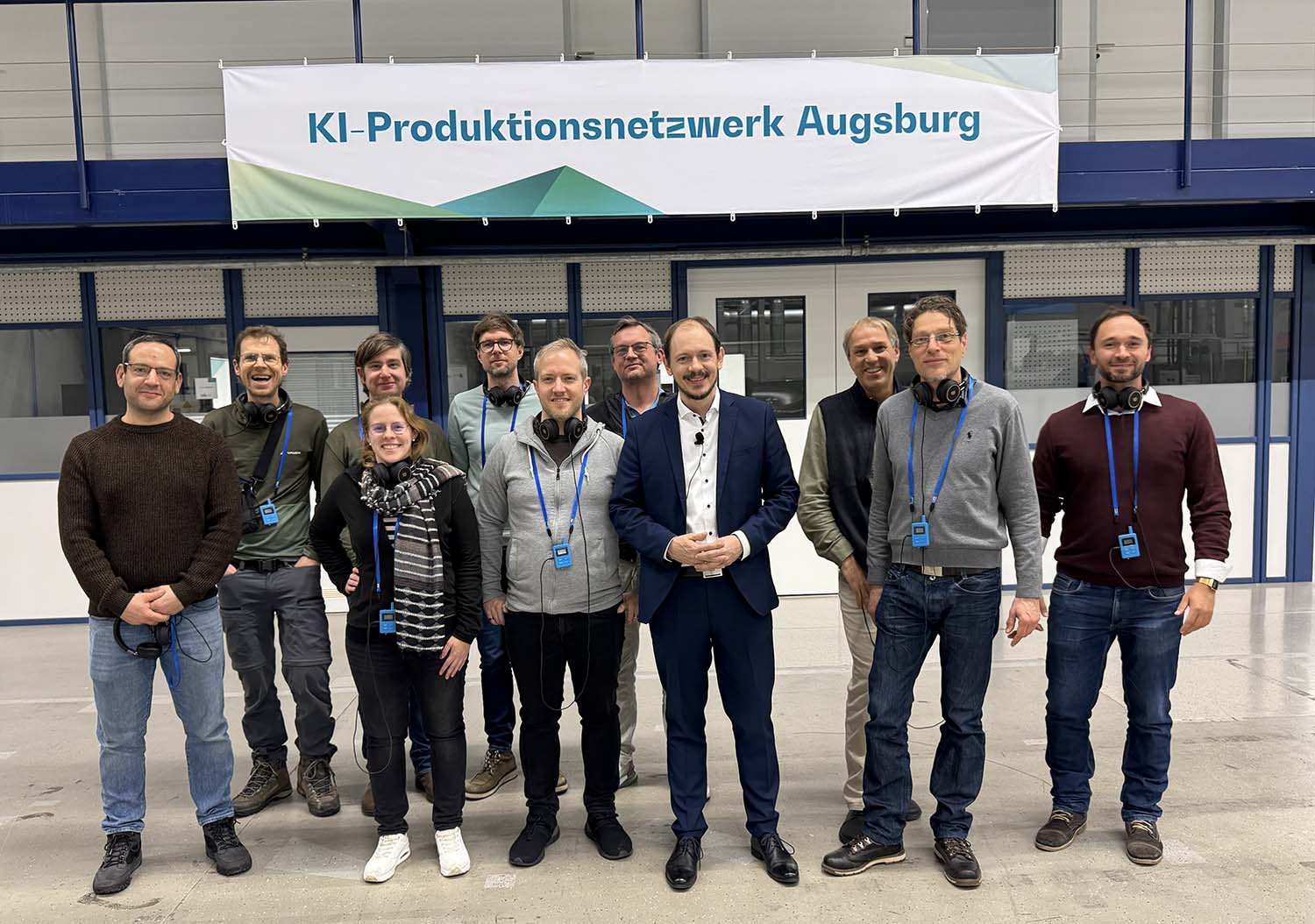 Gruppenfoto der Teilnehmenden der DFN-Cloud Unkonferenz 2026 in einer Industriehalle der Universität Augsburg. Im Hintergrund hängt ein Banner des KI-Produktionsnetzwerks Augsburg. Foto: Dirk Bei der Kellen, DFN-Verein