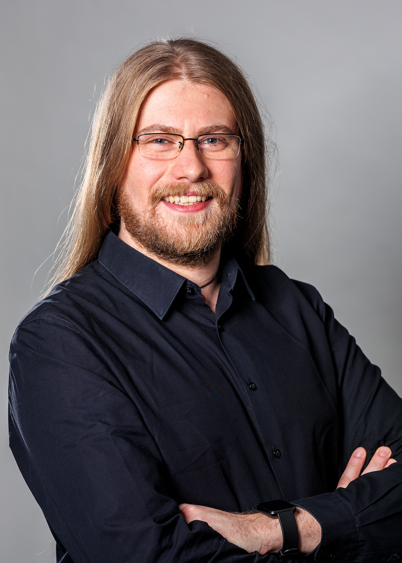 Ein professionelles Porträtfoto eines Mannes mit langem, hellem, glattem Haar, einem dichten Bart und Brille. Er lächelt freundlich und steht in einem schwarzen Hemd mit verschränkten Armen vor einem neutralen, grauen Hintergrund. An seinem Handgelenk trägt er eine schwarze Smartwatch. Das Foto ist scharf und gut ausgeleuchtet.